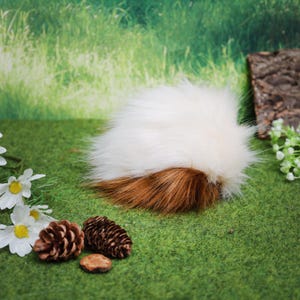May include: A fluffy, two-toned faux fur pompom, white on top and brown at the base. The pompom is set on a green surface with decorative elements like pinecones and flowers, suggesting a craft or accessory item.