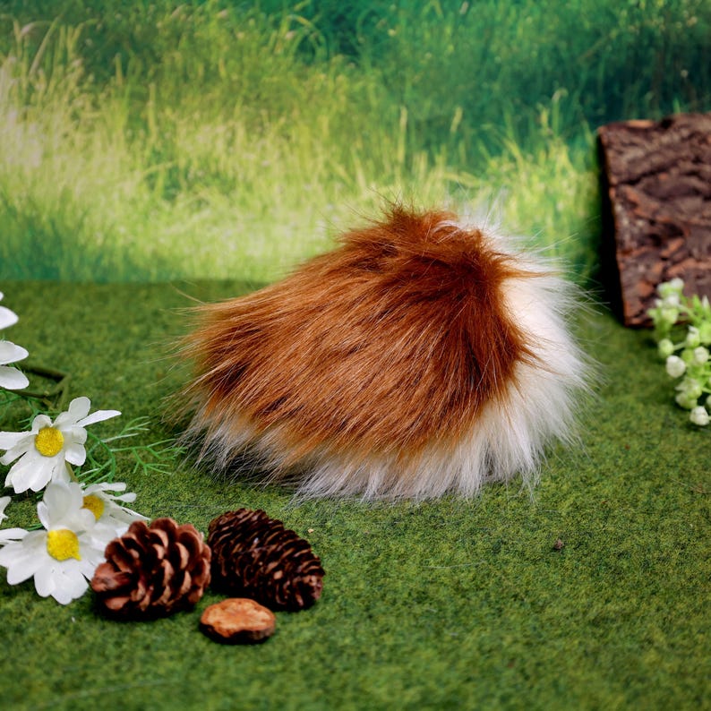 May include: A fluffy, faux fur tail in shades of brown and white, with a rounded shape. The tail is set against a green background with decorative elements like pinecones and flowers, suggesting a woodland theme.