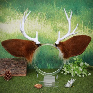 May include: A headband featuring brown faux fur deer ears and white antlers. The ears are attached to a clear plastic headband. The antlers are white and have a realistic appearance. Perfect for cosplay or themed events.