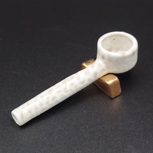 Puede incluir: Un cucharón de cerámica blanca con un acabado moteado. El cucharón tiene un mango largo y un cuenco redondeado. Está apoyado en un soporte dorado.