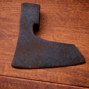 Op de afbeelding: Een verweerde bijlkop met een donkere, roestige afwerking. De metalen bijlkop heeft een gebogen blad. Het ligt op een houten oppervlak met een warme, bruine tint.