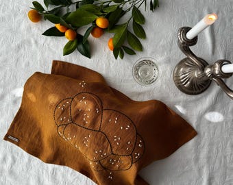 May include: A brown linen tea towel with an embroidered challah bread design. The towel is folded and lying on a white surface with a silver candlestick and citrus fruit in the background.
