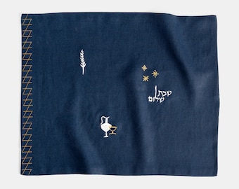 May include: A navy blue rectangular textile with gold stitching along the left edge. The textile features embroidered white and gold designs, including a wheat stalk, stars, a pitcher, a glass, and Hebrew text.