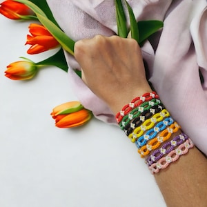 May include: A stack of colorful friendship bracelets with daisy flower accents. The bracelets are made of woven string and feature a variety of colors, including red, orange, yellow, green, blue, purple, and pink.