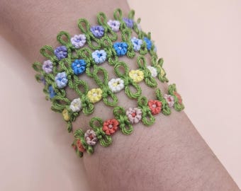 May include: A set of four crocheted bracelets with green string and colorful flower accents. The bracelets are stacked on a person's wrist.