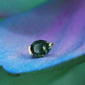 May include: A teardrop-shaped, dark green gemstone set in a gold-colored metal. The jewelry piece is displayed on a blue and purple petal. The text "BROWNCOAT PIERCER" is at the bottom of the image.