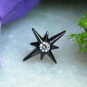 May include: A black metal starburst-shaped body jewelry piece featuring a single clear round gemstone at its center. The design includes eight pointed rays of varying lengths radiating from the middle. The text BROWNCOAT PIERCER is visible.