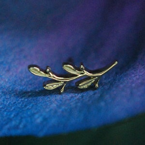 May include: A pair of gold-colored leaf-shaped earrings with small, sparkling accents. The earrings are set against a dark blue and purple background. The text "BROWNCOAT PIERCER" is at the bottom of the image.