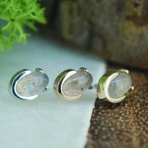 May include: Three silver and gold body jewelry pieces with a crescent moon design and a light gray gemstone. The jewelry is on a white surface.