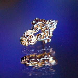 May include: A gold-colored ornate jewelry piece with a swirling design, reflecting on a blue surface. The piece is likely a body piercing.