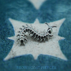May include: A silver-coloured, articulated centipede-shaped body jewellery piece. The jewellery is highly detailed with multiple segments and small legs. The piece is displayed on a textured, light-coloured surface with a blue starburst pattern in the background. The text "BROWNCOAT PIERCER" is visible.