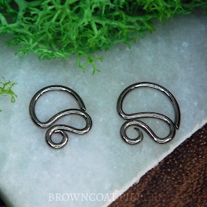 Könnte beinhalten: Zwei dunkelmetallische, schusswaffengraue Piercingringe mit geschwungenem, wirbelndem Design. Jedes Stück besteht aus einem kreisförmigen Ringsegment, das in eine S-förmige dekorative Verzierung übergeht und eine durchgehende, fließende Metalldrahtform bildet.