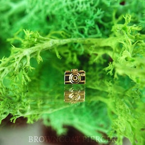 May include: A gold-colored camera charm with a black lens. The charm is sitting on a green surface.