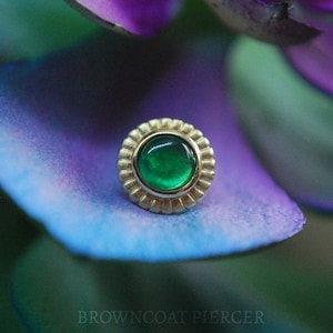 May include: A green gemstone set in a gold bezel with a decorative border. The gemstone is surrounded by a gold setting with a textured edge.
