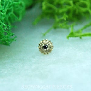 May include: A gold-colored, ornate stud earring with a central black stone. The earring has a circular design with detailed patterns around the stone. The text "BROWNCOAT PIERCER" is visible in the background.
