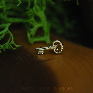 May include: A gold-toned key charm with an ornate design. The key is lying on a brown wooden surface.