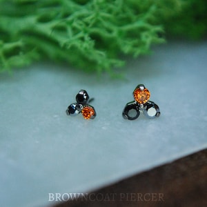 May include: Two pieces of jewelry, each with a cluster of three gemstones. The gemstones are black, orange, and silver. The jewelry is set against a white surface with green foliage in the background. The text "BROWNCOAT PIERCER" is at the bottom.