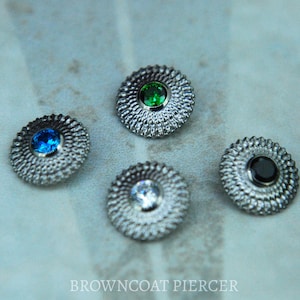 Op de afbeelding: Vier zilverkleurige, getextureerde, ronde sieraden met een centrale edelsteen. De edelstenen zijn blauw, groen, helder en zwart. De tekst "BROWNCOAT PIERCER" staat onderaan de afbeelding.
