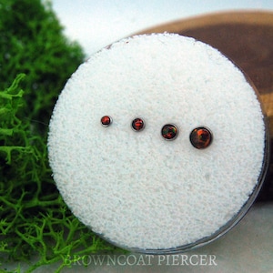 May include: Four circular stud earrings arranged in a row by size, each featuring a dark, iridescent stone with flecks of orange and red colour set in a metallic bezel. The studs are displayed on a white, textured circular foam pad.