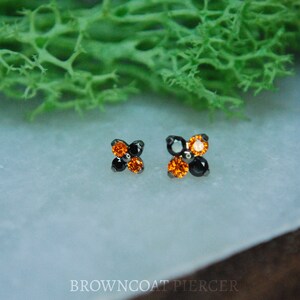 May include: Two small, black, four-pronged, prong-set, body jewelry pieces with orange and black gemstones.