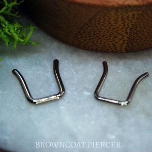 May include: Two silver U-shaped nose rings, also known as a septum ring, on a white surface.  BROWNCOAT PIERCER is printed in black text below the rings.