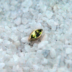 May include: A marquise-shaped, olive-green gemstone set in a gold bezel. The gemstone is surrounded by a gold setting and rests on a bed of white, granular material. The image is a close-up shot, highlighting the details of the jewelry.