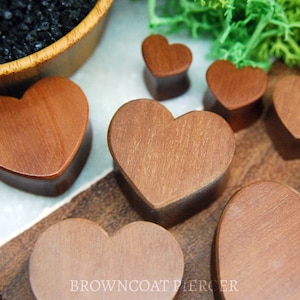 May include: A collection of heart-shaped wooden plugs in shades of brown. The plugs vary in size and display a natural wood grain. The image also shows a wooden bowl with black stones and green foliage. The text "BROWNCOAT PIERCER" is visible.