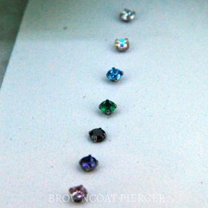 May include: A selection of small, colorful gemstone studs arranged in a vertical line. The gems include clear, iridescent, blue, green, black, purple, and pink. The text "BROWNCOAT PIERCER" is visible at the bottom.