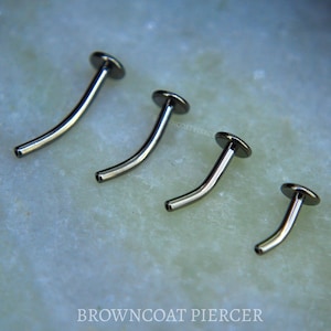 May include: Four curved barbell body jewelry pieces in a dark metallic finish, arranged in descending order of length. Each piece features a flat circular disc at one end and a hollow opening at the other. The text BROWNCOAT PIERCER is visible.