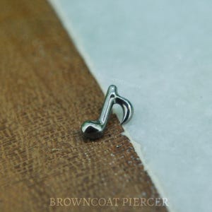 Pode incluir: Uma pequena nota musical prateada. A joia está colocada sobre uma superfície de madeira castanha texturizada e um fundo azul claro. O texto "BROWNCOAT PIERCER" é visível na imagem.