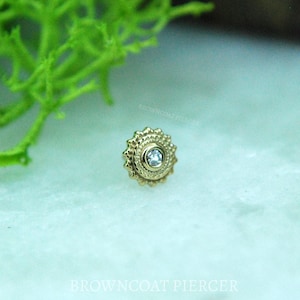 May include: A gold-colored, ornate stud earring with a central, clear gemstone. The earring has a sunburst design with a textured border. The background is a light, neutral color with a hint of green.