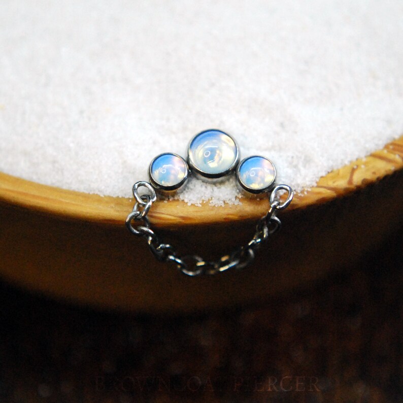 Pode incluir: Uma corrente de prata com tr&ecirc;s pedras opalescentes, cada uma com uma cor iridescente diferente, dispostas em uma linha curva. A corrente est&aacute; sobre uma superf&iacute;cie branca.