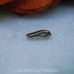 May include: A silver coffin-shaped body jewelry piece with a cross engraved on the top.