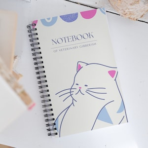 May include: A white spiral bound notebook with a light blue and pink design. The cover features a cartoon cat with a blue and white collar and the text "NOTEBOOK OF VETERINARY GIBBERISH".