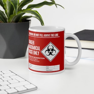 May include: White ceramic mug with a red and black biohazard warning label. The label reads "Warning Do Not Fill Above The Line", "Danger Anatomical Waste Only", "Destroy by Incineration or other Suitably Authorised Process", "Date", "Date", and "Dept/Ward".