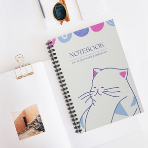 以下が含まれることがあります： ピンクの耳と青いアクセントが付いた漫画の猫が描かれた、薄い灰色の表紙のスパイラルバインディングノート。表紙には黒い文字で「NOTEBOOK OF VETERINARY GIBBERISH」と書かれています。