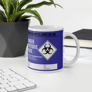 Peut inclure: Mug en céramique blanche avec une bande bleue portant les mots "DANGER CYTOTOXIC WASTE" et un symbole de risque biologique. La tasse comprend également le texte "WARNING DO NOT FILL ABOVE THE LINE".