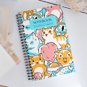 以下が含まれることがあります： 猫、魚、ハート、その他の動物関連のイラストが描かれた、青と白のスパイラルノート。表紙には「Notebook of Veterinary Gibberish」と書かれている。