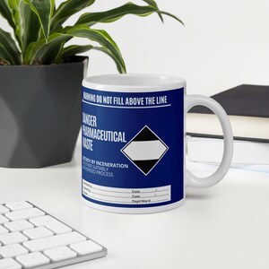 Peut inclure: Une tasse en céramique bleue avec une poignée blanche. La tasse comporte une étiquette d'avertissement qui indique "WARNING DO NOT FILL ABOVE THE LINE" et "DANGER PHARMACEUTICAL WASTE". L'étiquette comprend également un symbole en forme de losange noir et blanc et des instructions de mise au rebut.