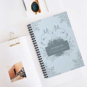 以下が含まれることがあります： 表紙に花柄と2匹の猫が描かれた青いスパイラルノート。表紙のテキストは「Notebook of Veterinary Gibberish」です。