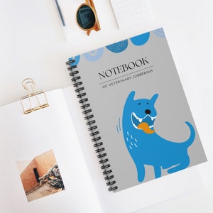 May include: A gray spiral bound notebook with a blue dog cartoon on the cover. The dog is holding a yellow fish in its mouth. The text on the cover reads "Notebook of Veterinary Gibberish".