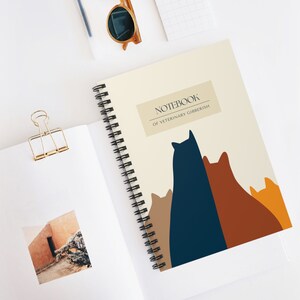 以下が含まれることがあります： ベージュの表紙に「Notebook of Veterinary Gibberish」という文字が書かれた、スパイラルバインディングされたノート。表紙には、茶色、オレンジ、青の様々な色合いの猫5匹のグラフィックが描かれている。