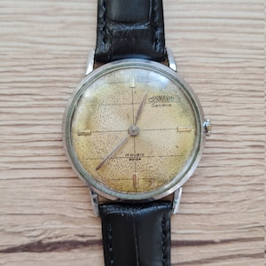 May include: A silver wristwatch with a black leather band. The watch face is yellowed and has black markings. The watch face says "CORNAVIN Geneve" and "17 RUBIS SWISS".