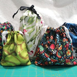 May include: Five drawstring bags with different patterns. The bags are made of fabric and have white drawstrings. The patterns include a green and white vegetable pattern, a green and red baseball pattern, a black and colorful floral pattern, a blue and white star pattern, and a black and red fruit pattern.