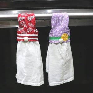 May include: Two hanging kitchen towels. One towel has a red top with the word "Indiana" in white script. The other towel has a purple floral top with a green band and yellow flower accent. Both have white towels.