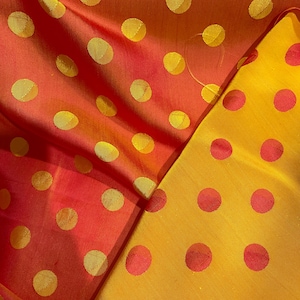 Könnte beinhalten: Nahaufnahme eines Stoffes mit Polka-Dot-Muster. Der Stoff ist eine Mischung aus Orange- und Gelbtönen mit großen, gleichmäßig verteilten Polka-Dots in kontrastierenden Farben. Der Stoff ist gefaltet und zeigt beide Seiten des Materials.