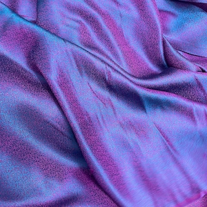 Puede incluir: Primer plano de una tela con superficie texturizada. La tela tiene un degradado de color púrpura, magenta y turquesa. La tela está drapeada, creando pliegues y sombras que añaden profundidad y dimensión a la imagen.