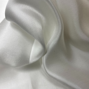 Könnte beinhalten: Nahaufnahme eines glatten, weißen Stoffes mit seidenartiger Textur. Der Stoff ist drapiert und gefaltet und erzeugt weiche Kurven und Falten.
