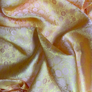Op de afbeelding: Close-up van een gevouwen stof met een bloemenpatroon. De stof is een mix van gele en roze tinten, met het bloemmotief in een lichtere roze tint. Het materiaal lijkt zijdeachtig en heeft een lichte glans.