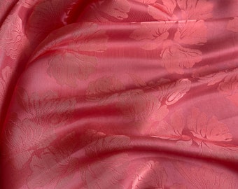May include: Close-up of a pink fabric with a floral pattern. The fabric has a satin sheen and is draped in soft folds, creating shadows and highlights. The floral design is subtly raised, adding texture to the material. The overall effect is elegant and luxurious.
